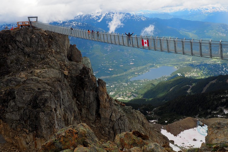 mountains and suspension bridge at the top of Whistler peak in summer months with canada flag and whistler village in the distance