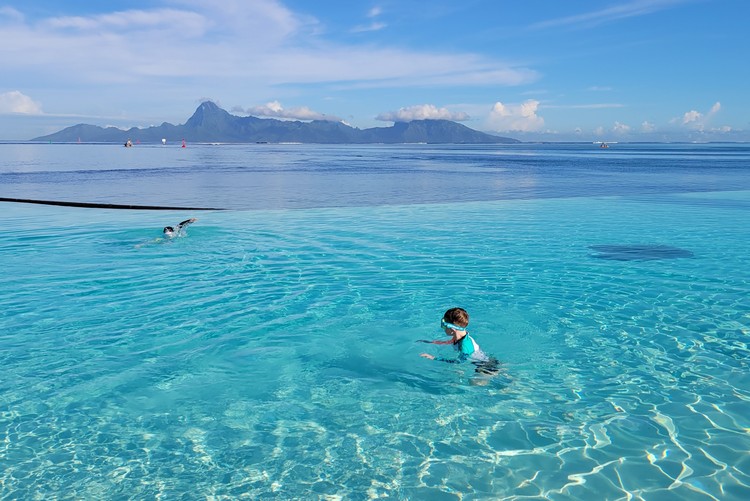 infinity pool at Te Moana Tahiti Resort with view of Moorea Island in the distance. Kids swimming in the pool with blue water